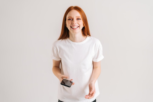 Studio portrait of cheerful young woman with wide smile reading good online message using mobile phone on white isolated background Happy lady holding smartphone in hands and looking at camera