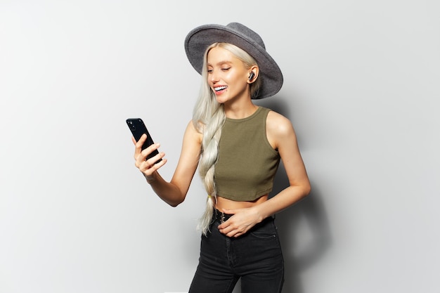 Studio portrait of cute happy blonde girl while dancing and listen the music via wireless earbuds holding smartphone in hand wearing grey hat on white background