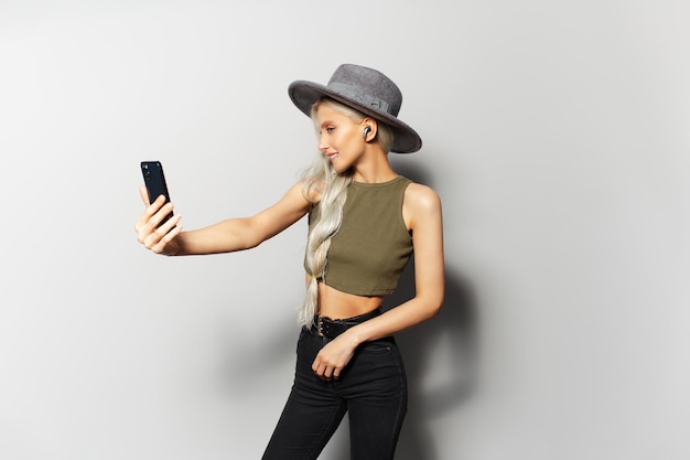 Studio portrait of cute happy blonde girl while making selfie photo and listen the music via wireless earbuds holding smartphone in hand wearing grey hat on white background