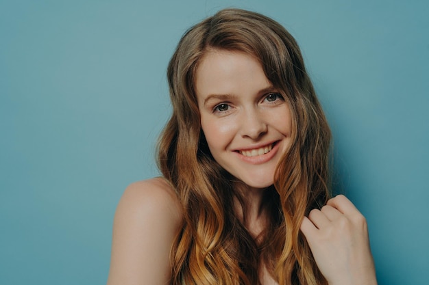 Studio portrait of cute teen girl with wavy hair smiling and looking at camera isolated on blue