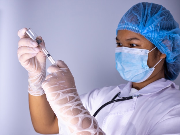 마스크를 쓰고 모자를 쓰고 있는 여성 의사의 스튜디오 초상화. 손에는 백신 한 병과 주사기 한 자루가 들려 있었다. 흰색 배경에 서. 스튜디오 촬영 배경, COVID-19 개념