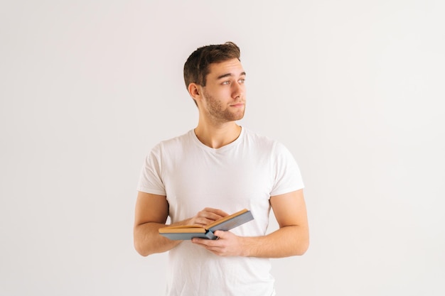 Студийный портрет сосредоточенного молодого человека, держащего в руках бумажную книгу на белом изолированном фоне в студии, отводящего взгляд. Вид спереди серьезного студента-мужчины, изучающего чтение учебных материалов.