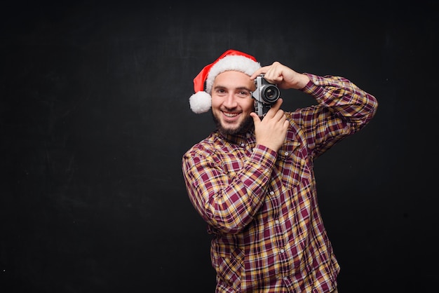 Ritratto dello studio dell'uomo barbuto divertente e sorpreso che porta il cappello della santa che tiene una retro macchina fotografica, facendo una foto. spazio per il testo. nero