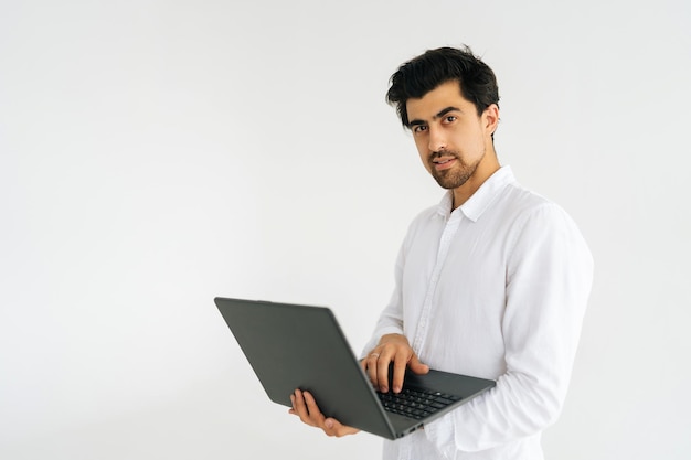 Studio portrait of handsome bearded young man posing with laptop computer and looking at camera standing on white isolated background