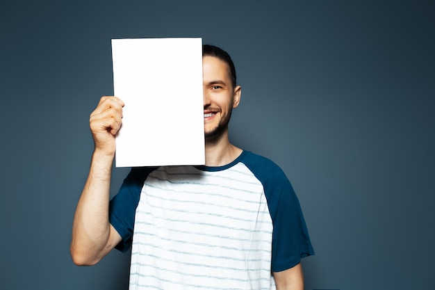 Studio portrait of handsome smiling guy holding white empty paper board on half face on blue background