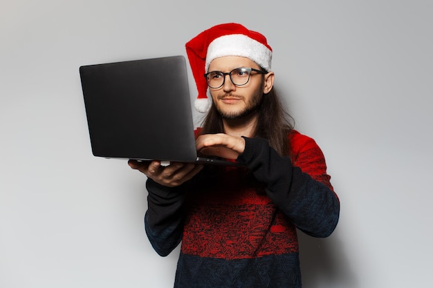 Studio portrait of handsome thoughtful guy holding laptop and typing on keyboard Wearing Santa Claus hat Christmas concept White background