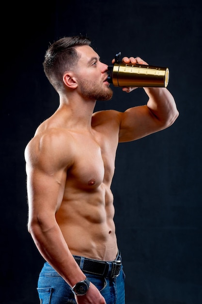 Studio portrait of handsome young bodybuilder holding bottle and drinking over black background Half naked athlete with strong body