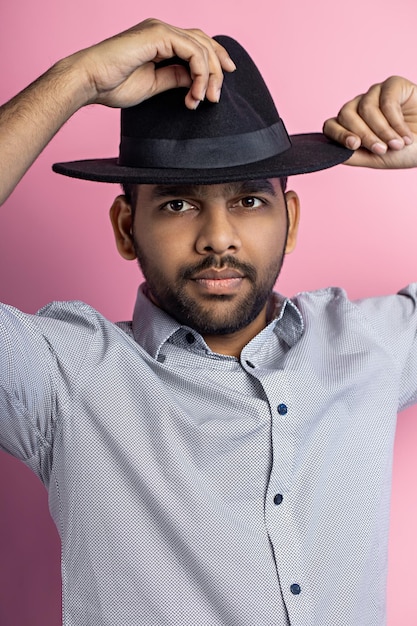 Photo studio portrait of handsome young indian male with small beard, wearing gray shirt and black hat, looking at camera with serious expression, posing over pink background.