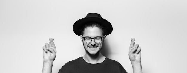 Studio portrait of happy guy crosses fingers with closed eyes on grey background Panoramic banner Black and white photo