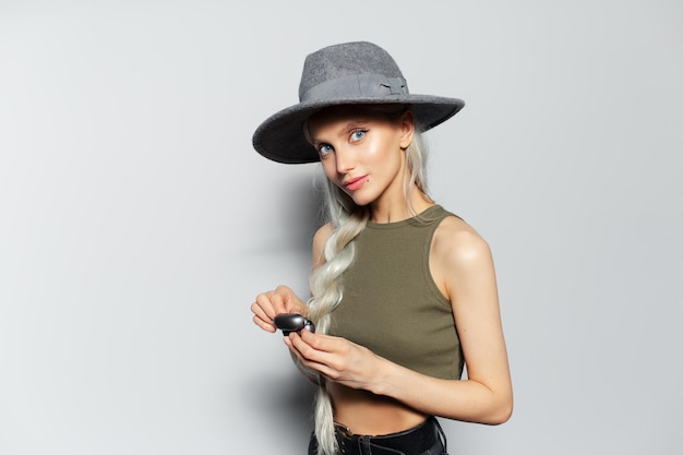 Studio portrait of happy pretty blonde girl holding charging case for earbuds in hands wearing grey hat on white background