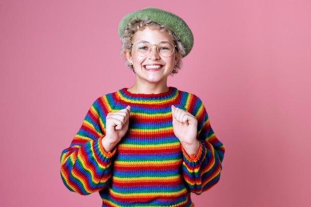 Ritratto in studio di una donna felice con un maglione arcobaleno e occhiali su una parete rosa