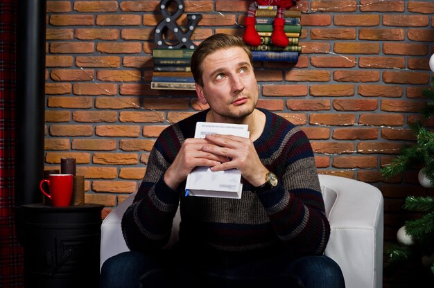 Ritratto in studio di uomo con libro seduto su una sedia contro l'albero di natale con decorazioni.