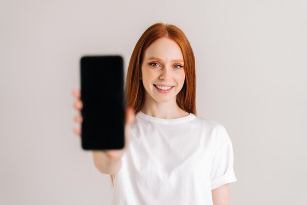 Фото Студийный портрет веселой рыжей молодой женщины с широкой улыбкой, показывающей пустой экран мобильного телефона
