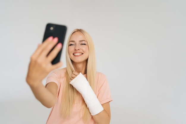 Studio portrait of positive young blonde woman with broken arm wrapped in plaster bandage talking smartphone making video call taking selfie picture