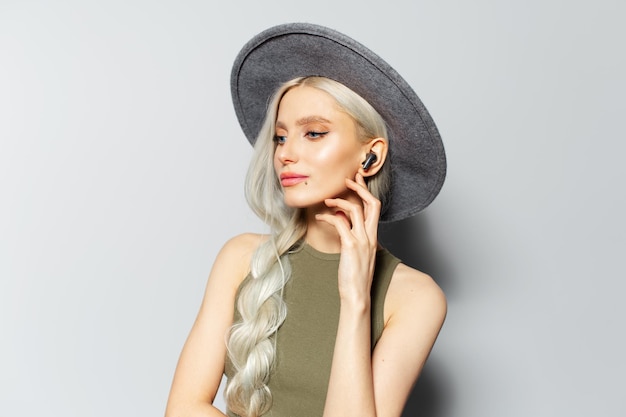 Studio portrait of pretty blonde girl touching wireless earbuds while listening to music on white background Wearing grey hat