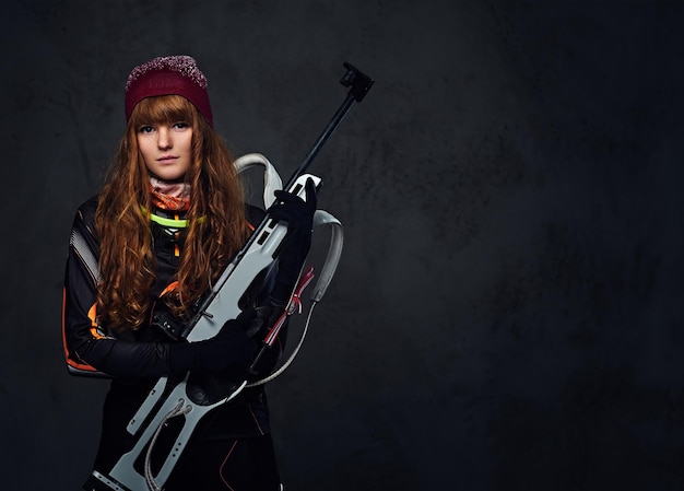 Studio portrait of a redhead female Biathlon champion holds a gun over grey background.