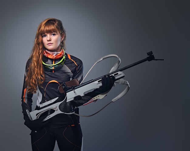 Ritratto in studio di una campionessa di biathlon rossa tiene una pistola su sfondo grigio.