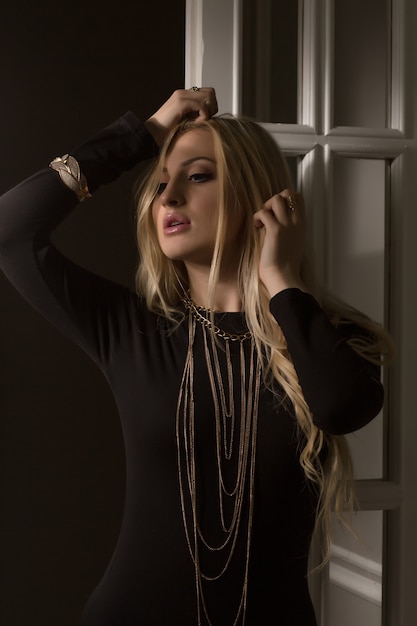 Студийный портрет чувственной блондинки-модели в черном платье с золотым ожерельем