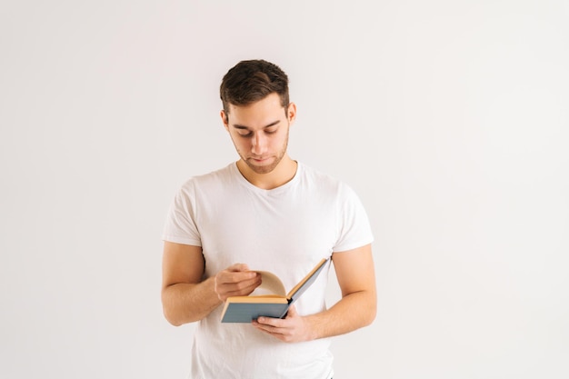 Studio portrait of serious young man turning pages of paper book on white isolated background in studio. Front view of handsome male student studying reading educational materials.