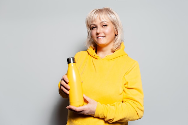 Studio portrait of smiling blonde woman holding metal thermo water bottle on white background Yellow color