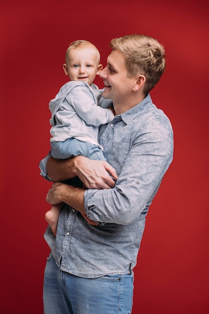 Studio portrait of smiling happy dad in denim shirt and jeans hugging his son in hands who is looking at camera. Isolated on red background. Father s day.