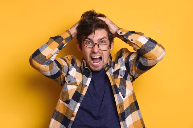 Studio portrait of stressed man in angry, experiencing stress, madness, he furiously rips out his hair and screams. Isolated on yellow background. Copy space. Concept of mental health, human emotions