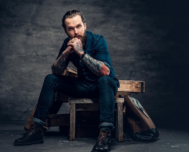 Studio portrait of stylish bearded male with tattoos on arms, sits on wooden boxes over grey background in a studio.