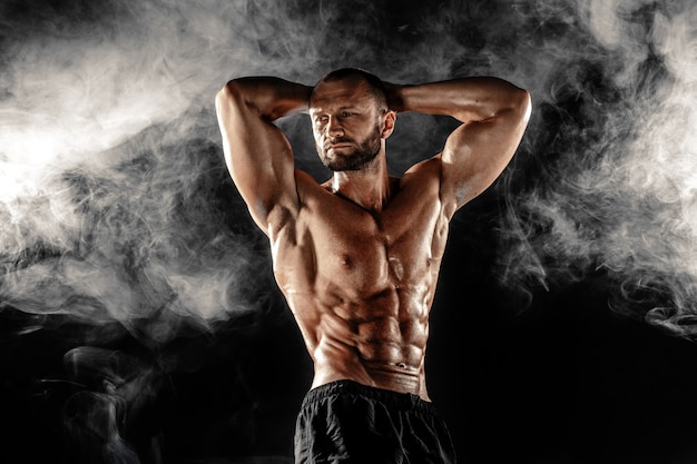 Studio portrait of topless muscular sportsman posing with arms on head over smoke black scene.