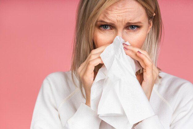 Studio portrait of an unhealthy pretty blonde woman in casual blowing her nose napkins, looking at the source of the allergy, rhinitis, cold, allergy concept isolated over pink background.