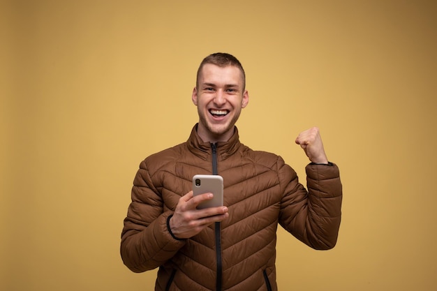 Studio portrait. Young 20s man in a brown jacket, on a yellow background, holding in his hand, uses a mobile phone, makes a winner gesture, clenching his fist, say "yes"