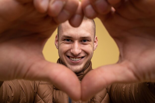 Photo studio portrait. young 20s man in brown jacket, on yellow background, showing heart shape, look through hands, heart shaped sign