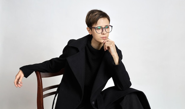 Studio portrait of a young attractive girl sitting on a chair in a black coat on a gray background.