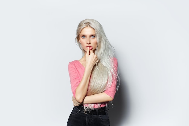 Studio portrait of young blonde girl touching lips wearing pastel pink shirt on white background