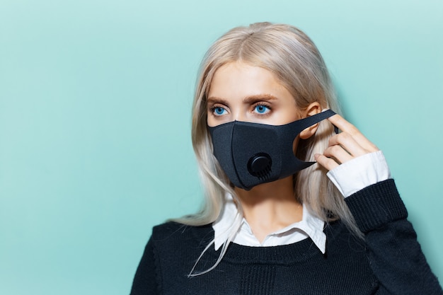 Photo studio portrait of young blonde girl with blue eyes in school form, wearing black respiratory face mask against coronavirus on background of cyan, aqua menthe color.