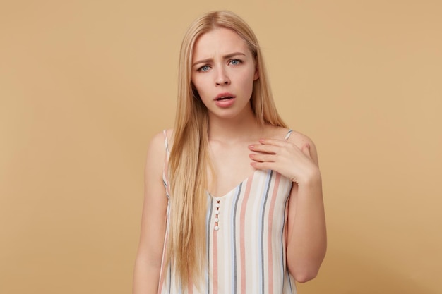 Studio portrait of young blonde woman posing over beige background looking into camera and points at her self feels guilty