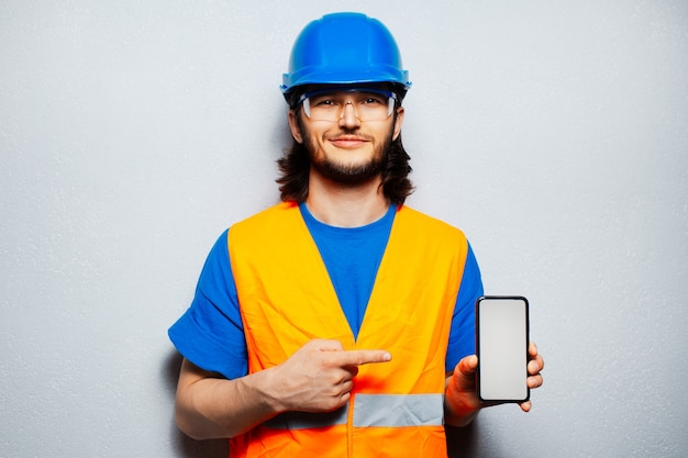 Studio portrait of young construction worker engineer wearing safety equipment, pointing finger at smartphone