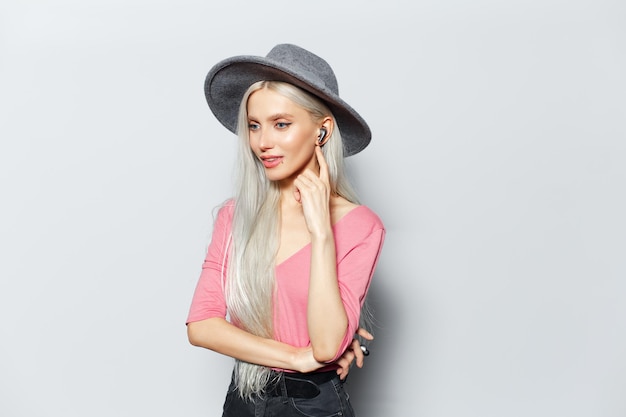 白い背景にグレーの帽子とピンクのシャツを着て、耳にワイヤレスイヤホンを持つ若いかわいいブロンドの女の子のスタジオポートレート