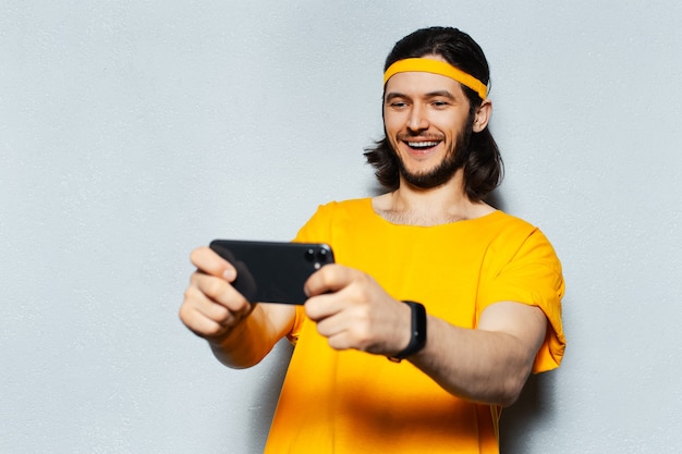 Studio portrait of young excited man playing game on smartphone, on grey textured background; wearing smartwatch, yellow band for head and shirt.