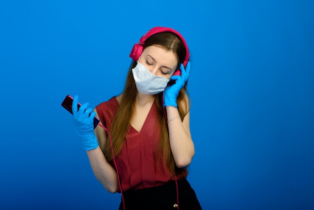 Studio portrait of young girl wearing medical flu mask and disposable gloves, female posing isolated over blue wall. Coronavirus, covid19.