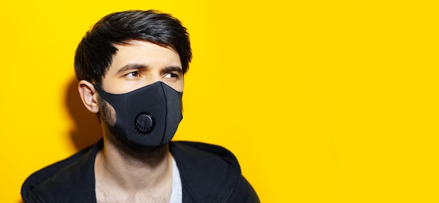 コピースペースと黄色の背景にコロナウイルスに対して黒い呼吸フェイスマスクを身に着けている若い男のスタジオの肖像画。