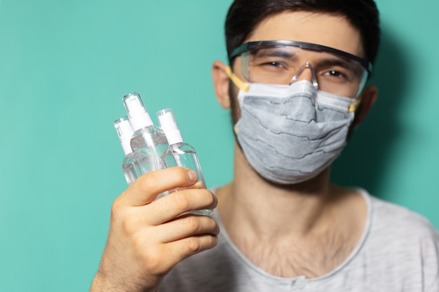 Studio portrait of young guy wearing medical flu mask and safety goggles against coronavirus, holding dispenser bottles with sanitizer antiseptic gel, on surface of cyan