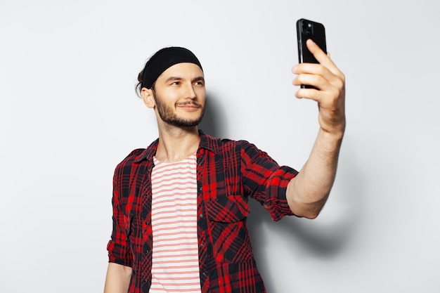 赤いカジュアルな服と黒いヘッドバンドを身に着けている白い背景の上のスマート フォンで selfie 写真を作る若いハンサムな笑みを浮かべて男のスタジオ ポートレート