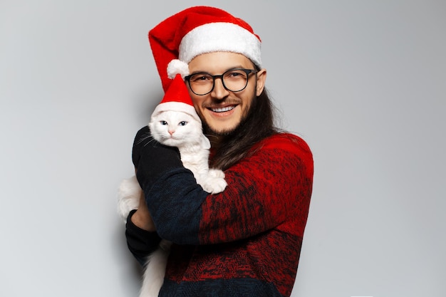 Studio portrait of young happiness man wears red Christmas sweater hugs a white cat wearing Santa hat on white background