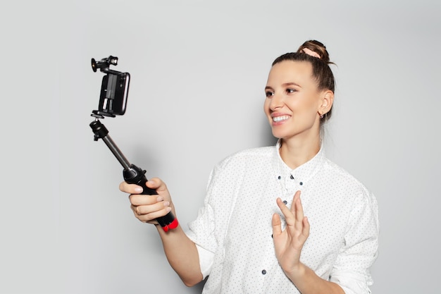 Ritratto in studio di giovane ragazza felice che fa selfie con lo smartphone sul bastone su sfondo grigio