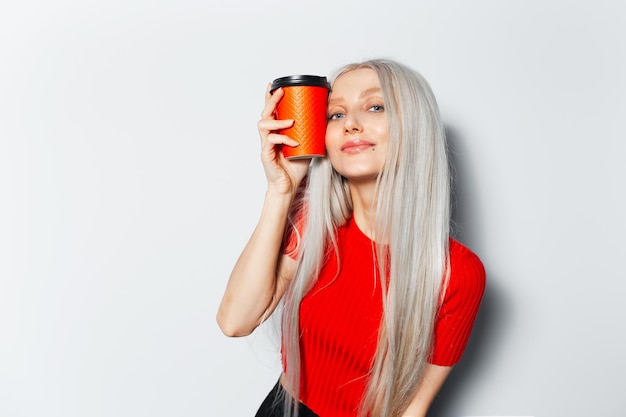 Studio portrait of young joyful pretty girl with blonde hair holding paper cup of coffee takeaway wearing red shirt on white background