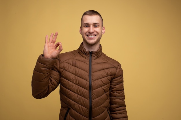 Studio portrait. A young man of the 20s in a brown jacket, on a yellow background, smiles and gestures that everything is fine