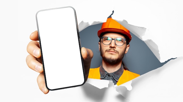 Studio portrait of young man construction worker showing big smartphone with blank on screen through torn paper hole White background Wearing eyeglasses and safety equipment