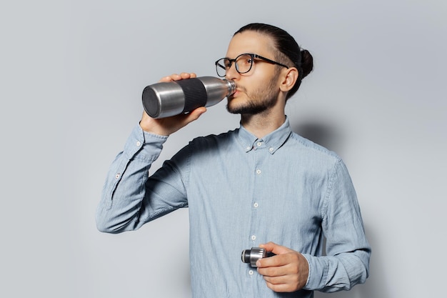 Ritratto in studio di giovane uomo che beve acqua da una bottiglia di metallo su sfondo bianco con occhiali da vista e camicia blu