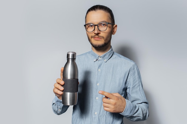 Studio portrait of young man pointing finger on metal bottle in his hand on white background wearing eyeglasses and blue shirt