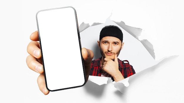 Studio portrait of young man, showing big smartphone with blank on screen through torn paper hole. W